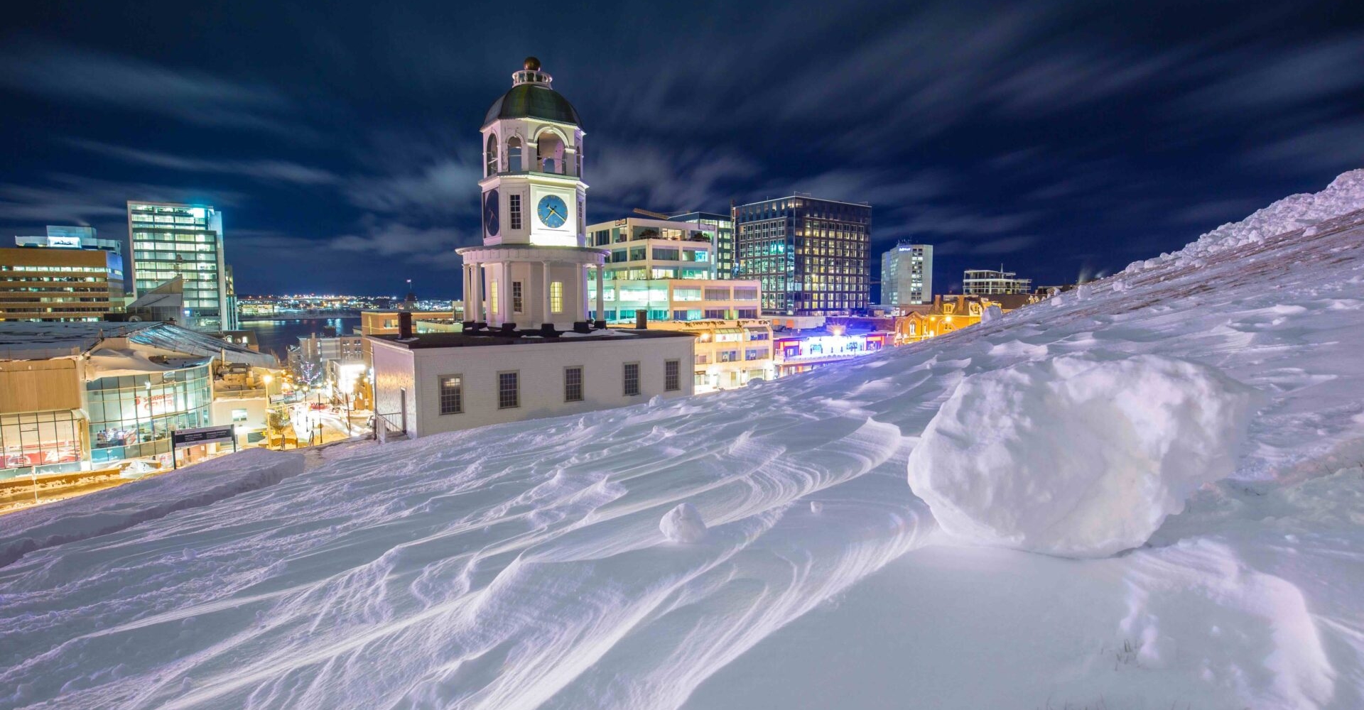 Picture of downtown halifax at night taken from Citadel hill. The town clock is in the foreground, with the hill covered in blown snow.